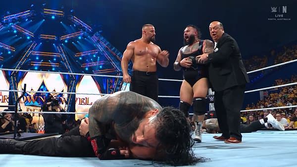 At WWE Crown Jewel in Perth, Roman Reigns lies defeated in the wrestling ring, while Bronson Reed celebrates alongside Bron Breakker and Paul Heyman. The background features vibrant lighting and a large logo for the event.