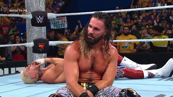 A victorious Seth Rollins, sitting on the wrestling mat, looks on as Cody Rhodes lies defeated on the ground after being knocked out. The crowd is visible in the background, with a mix of fans holding signs and wearing WWE merchandise.