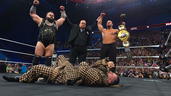 Seth Rollins lies defeated on the mat while his former stable celebrates above him, raising their arms in triumph. The atmosphere is charged with the energy of a WWE event.