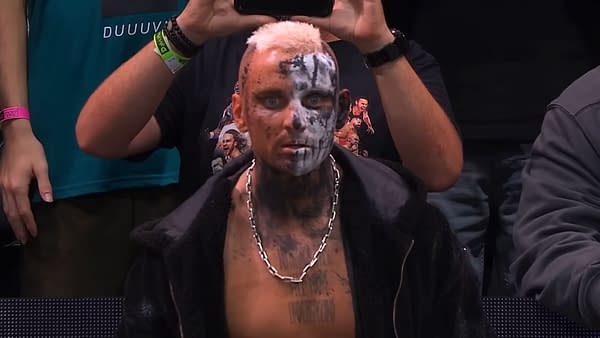 Darby Allin, a professional wrestler, shows off a striking look with half of his face painted in black and white and wearing a silver chain. He sits in the audience, visibly focused, with fans in the background.