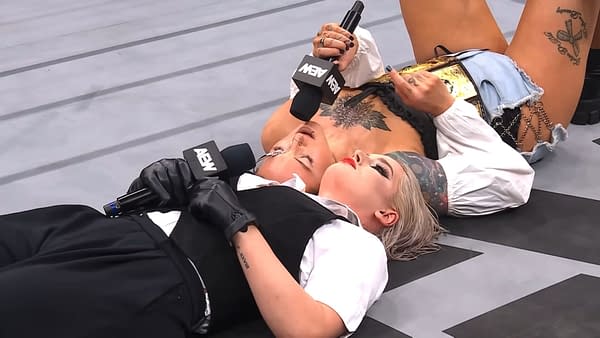Toni Storm and Kris Statlander chat while lying on the wrestling ring floor, each holding a microphone. Statlander has a championship belt next to her.
