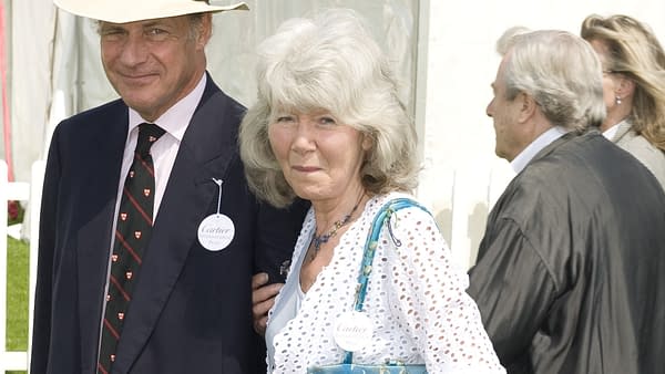 Dame Jilly Cooper, a notable author, arrives at the Cartier International Polo event in Windsor, wearing a white patterned dress and holding a blue handbag. She is accompanied by a man in a suit and hat, both sporting event badges.