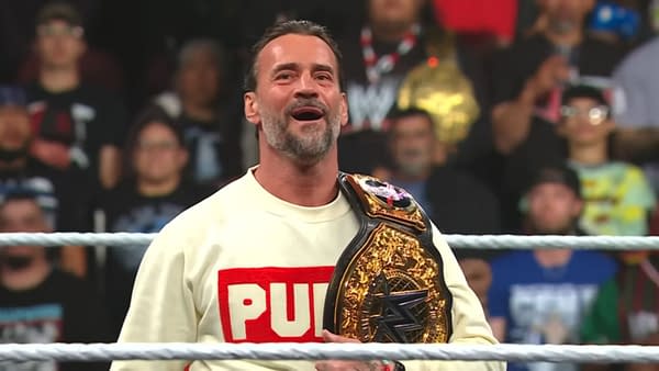 CM Punk stands in a wrestling ring wearing a white sweater with 'PUNK' printed on it and holding a championship belt, looking happy and relaxed. The audience is visible in the background, showing excitement.