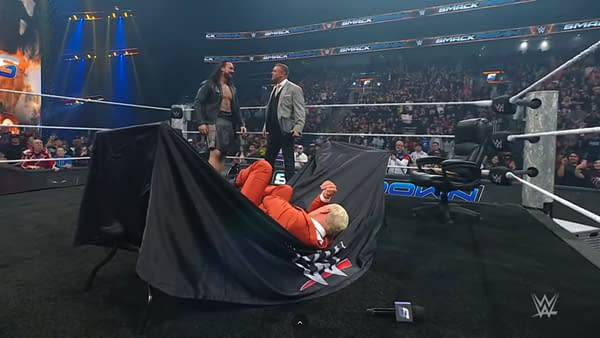 Cody Rhodes lies on the floor, partially covered by a black cloth, after a table break during a WWE SmackDown contract signing with Drew McIntyre. A referee and other officials stand nearby, surrounded by a cheering crowd.