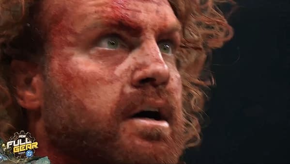 A close-up of a man with curly red hair and a beard, showing a shocked and distressed expression, covered in blood, likely reacting to a recent event in a wrestling match.