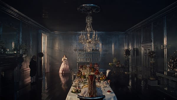 A lavish dining room from "Wuthering Heights" featuring a long table adorned with extravagant dishes and desserts, illuminated by a grand chandelier. In the background, a woman in a pink dress stands near a doorway, while a servant in formal attire silently observes.
