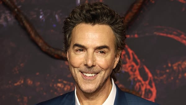 A smiling man with curly hair and a well-groomed beard is dressed in a blue suit, posing at what appears to be a promotional event related to Star Wars. The background features dark tones with red accents, suggesting a dramatic atmosphere.