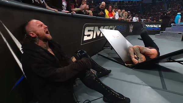 Aleister Black sits on the floor, visibly in pain, next to a Slim Jim-themed table that is broken and positioned upside down, with Damian Priest lying on the ground beneath it, during a WWE SmackDown event.