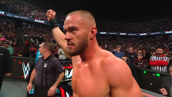 Austin Theory, a muscular wrestler with a crew cut, raises his fist in celebration after a match on WWE Raw, surrounded by a cheering crowd and staff members. The venue is packed with fans, creating an energetic atmosphere.