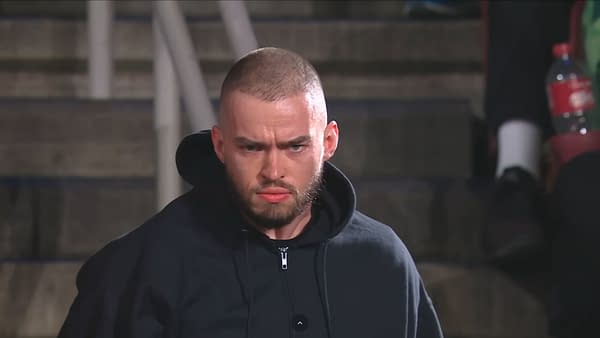 Austin Theory stands with a serious expression, wearing a black hoodie, as he unmasks during WWE Raw. The background features blurred audience members and stairs.