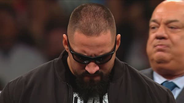 Bron Breakker, wearing sunglasses and a dark jacket, appears focused and prepared for an upcoming confrontation, with his head down and a serious expression. Paul Heyman can be seen in the background, watching him intently.