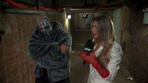 A wrestler with distinctive face paint and a furry coat is interviewed backstage in a dimly lit corridor, while a blonde woman in a white outfit speaks into a microphone. The background shows exposed brick and some debris on the floor.