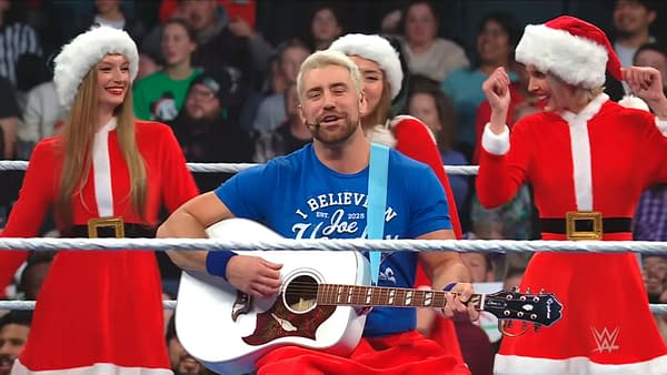 Joe Hendry, wearing a blue t-shirt, sings while holding an acoustic guitar, surrounded by women in Santa Claus outfits. The scene takes place inside a wrestling ring, with fans visible in the background.
