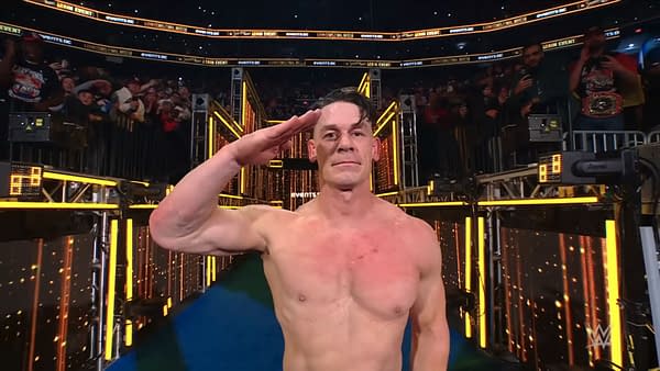 John Cena salutes the fans with a serious expression while shirtless, standing on a brightly lit stage following his last match at Saturday Night's Main Event. A crowd is visible in the background, cheering him on.
