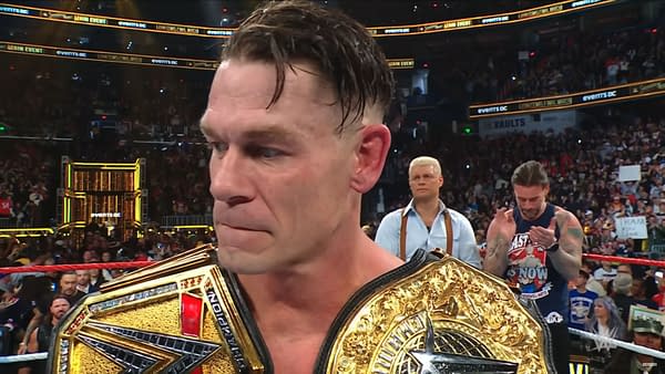 John Cena, holding two championship belts, expresses emotion as he stands amidst a cheering audience following his final match at Saturday Night's Main Event. Peers in the background show their support while the crowd is visibly excited.