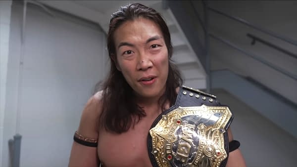 Konosuke Takeshita, a professional wrestler, is backstage holding a championship belt and speaks into the camera. He has long hair and appears engaged and determined.