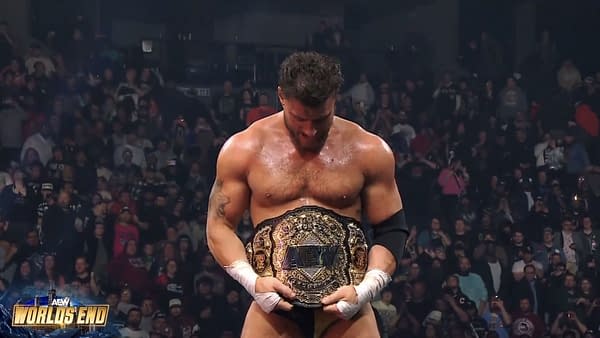A wrestler stands in the ring, holding the AEW World Championship belt and looking down at it, surrounded by an excited crowd of fans. The atmosphere is electric, reflecting the significance of the moment.