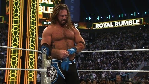 A wrestler with long hair, wearing a blue and black outfit, is taking off his blue gloves inside a wrestling ring after losing a retirement match. The crowd is visible in the background, with dim lighting and a large 'Royal Rumble' sign overhead.