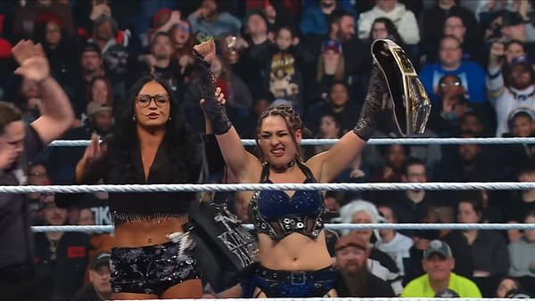 A female wrestler celebrates her victory by raising the US Title above her head in a WWE SmackDown wrestling ring. Another woman stands beside her, clapping in support, as fans cheer in the background.
