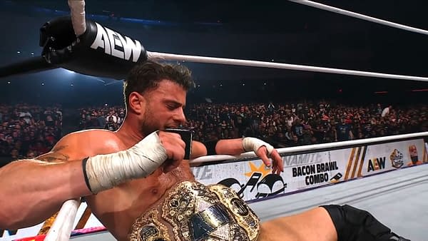 A visibly exhausted wrestler rests in the ring after a title defense at AEW Dynamite, gripping his championship belt while seated on the mat. The large crowd is visible in the background, capturing the atmosphere of the event.