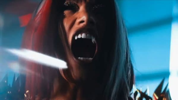A woman with long hair and dramatic makeup is passionately shouting during a promo video, conveying intense emotions. The vibrant background adds to the dynamic atmosphere.