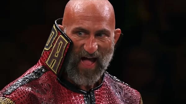 A bald, bearded wrestler wearing a red and black outfit with a high collar has a fierce expression on his face. His blue eyes are wide open, conveying intensity.