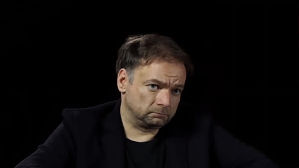 A middle-aged man with short, slightly tousled hair and a serious expression is seated against a black background. He wears a dark jacket over a black shirt, conveying a contemplative mood.
