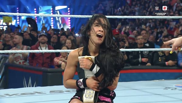 AJ Lee joyfully celebrates in the wrestling ring, holding the Intercontinental Championship belt close to her body after defeating Becky Lynch at the Elimination Chamber event, with an enthusiastic crowd cheering in the background.