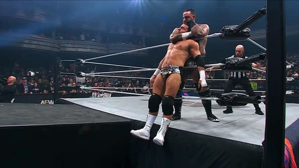 Brody King applies a chokehold to MJF, who is teetering at the edge of the wrestling ring, with referees and spectators visible in the background. The atmosphere is tense and electrifying.