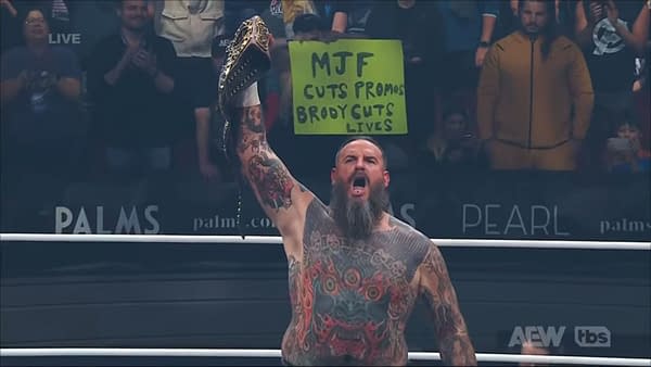 Brody King, a heavily tattooed wrestler with a beard, raises the AEW World Championship belt triumphantly in the ring, while shouting to the crowd. In the background, a fan holds up a sign that critiques wrestler MJF.