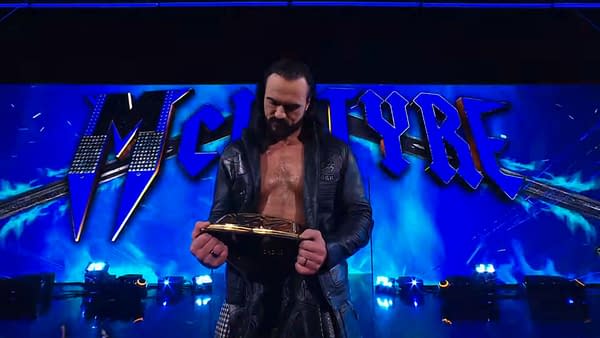 Drew McIntyre stands on stage, holding his championship belt and looking down at it. The background features blue lights and the name 'McIntyre' displayed prominently.