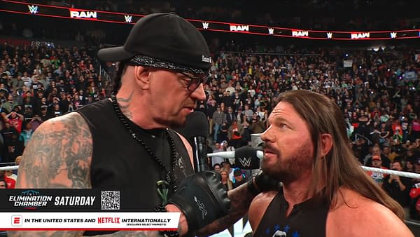 The Undertaker, wearing a black bandana and glasses, speaks into a microphone, announcing his WWE Hall of Fame induction to AJ Styles, who looks surprised. The audience is visible in the background, filling the arena with anticipation.