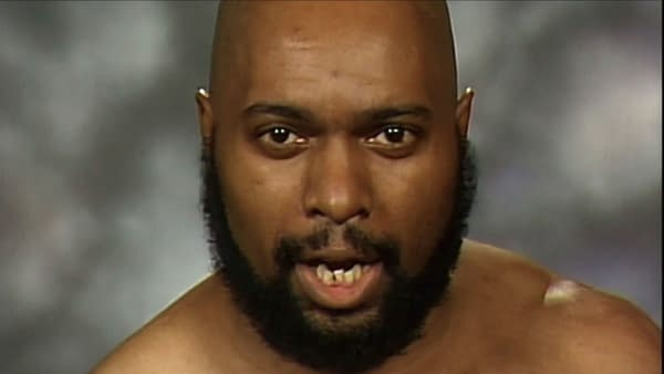 A close-up of a bald man with a beard, showing an intense expression. He has noticeable missing teeth and is dressed casually, against a blurred background.