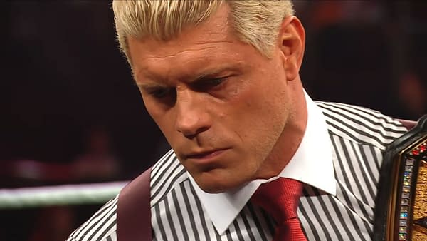 Cody Rhodes appears contemplative in the wrestling ring, wearing a striped shirt and red tie while holding a championship belt. He looks serious, addressing emotional themes.