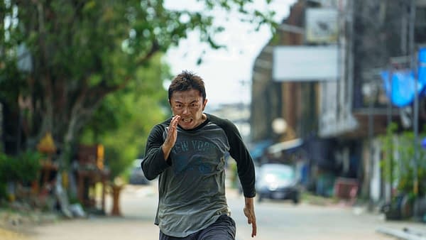 A man with a determined expression runs swiftly down a deserted street, wearing a grey and black shirt and grey shorts. The background features greenery and blurred structures, suggesting a tense moment.