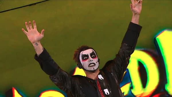 A performer in a dramatic costume, featuring bold face paint and a black jacket, raises both hands in the air during a WWE Raw event. The performer has a striking facial expression that adds to the comedic effect.