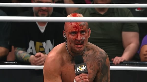 Darby Allin, showing signs of a brutal match with blood on his face and a determined expression, holds a microphone while addressing the audience on AEW Dynamite. He has visible tattoos, a chain around his neck, and a mohawk hairstyle.