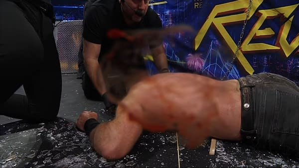 A wrestler lies face down in broken glass, with visible blood on their body, and a blood-covered camera lens in the foreground, capturing the aftermath of a violent Texas Death Match at AEW Revolution.