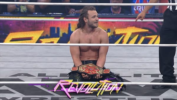 Jack Perry, visibly emotional and shirtless, holds up his newly won AEW National Championship belt while seated in the wrestling ring, surrounded by cheering fans and a vibrant background.