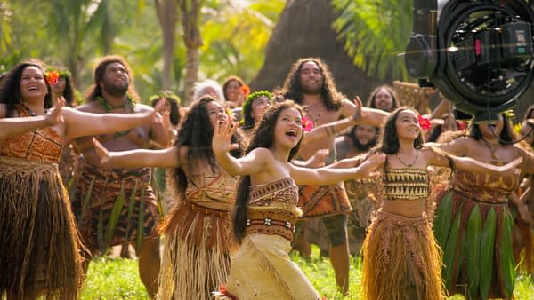MOANA A vibrant behind-the-scenes scene from Disney's live-action 'Moana,' featuring a group of performers dressed in traditional Polynesian costumes joyfully dancing in a lush outdoor setting. The image captures their enthusiastic expressions and dynamic movements, showcasing the cultural celebration central to the film.