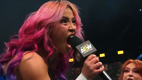Mina Shirakawa, with vibrant pink hair, passionately challenges her opponent on stage, holding a microphone labeled with the AEW logo.