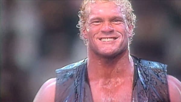 Sycho Sid Eudy, a wrestler with curly blonde hair and a wide smile, wearing a black vest, stands confidently in front of a blurred background. The wrestler appears sweaty and is showcasing a victorious expression.