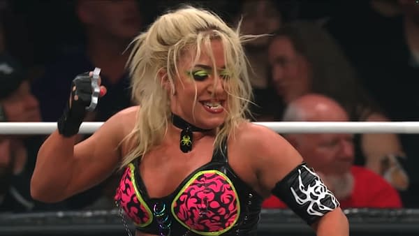 A female wrestler, Thekla, is in the ring during a match on AEW Dynamite, raising her fist with brass knuckles visible. She has long blonde hair, dramatic makeup, and wears a colorful outfit with a choker necklace.