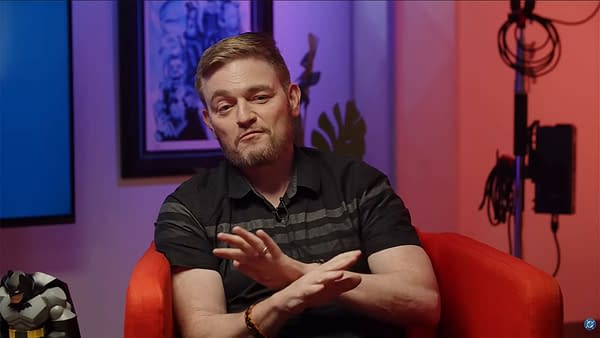 Scott Snyder DC Comics interview. A Batman figure is visible beside him, and colorful lighting adds a dynamic atmosphere.