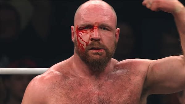 A muscular man with a bald head and beard displays a bloodied face, raising his arm triumphantly after a wrestling match. The marks of his battle are evident on his body.