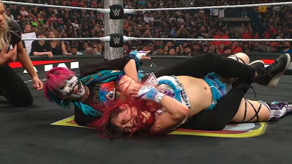 Asuka attacks Iyo Sky on WWE Raw, holding her down on the mat during a chaotic ringside brawl. A packed crowd watches as other wrestlers scramble nearby.