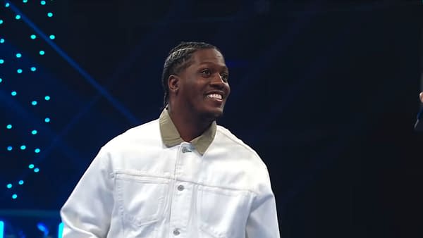 Lil Yachty smiles under blue stage lights on WWE SmackDown, wearing a white jacket.