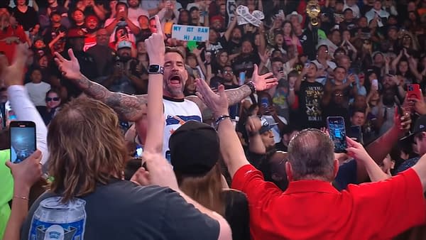 A man with tattoos and a tank top stands in a crowd, arms outstretched in celebration as fans cheer. The audience holds phones to capture the moment, with signs and various reactions visible throughout the crowd.