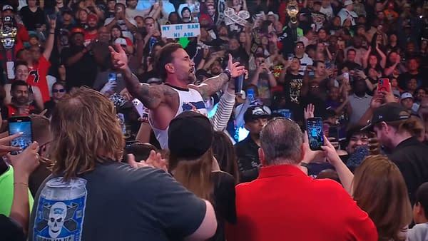 CM Punk stands in the center of a lively crowd during WWE Raw, arms open in celebration as fans cheer energetically. Many audience members are holding their phones to capture the moment.