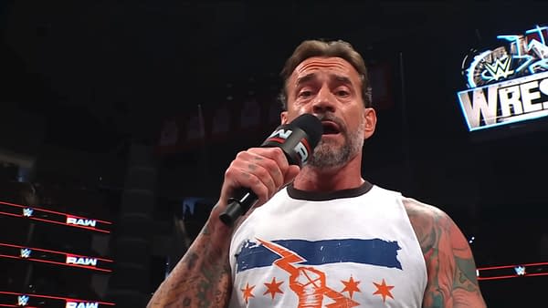 CM Punk passionately speaks into a microphone in an arena, with a WrestleMania sign illuminated in the background. He is wearing a white shirt with a blue and orange design and has visible tattoos on his arms.